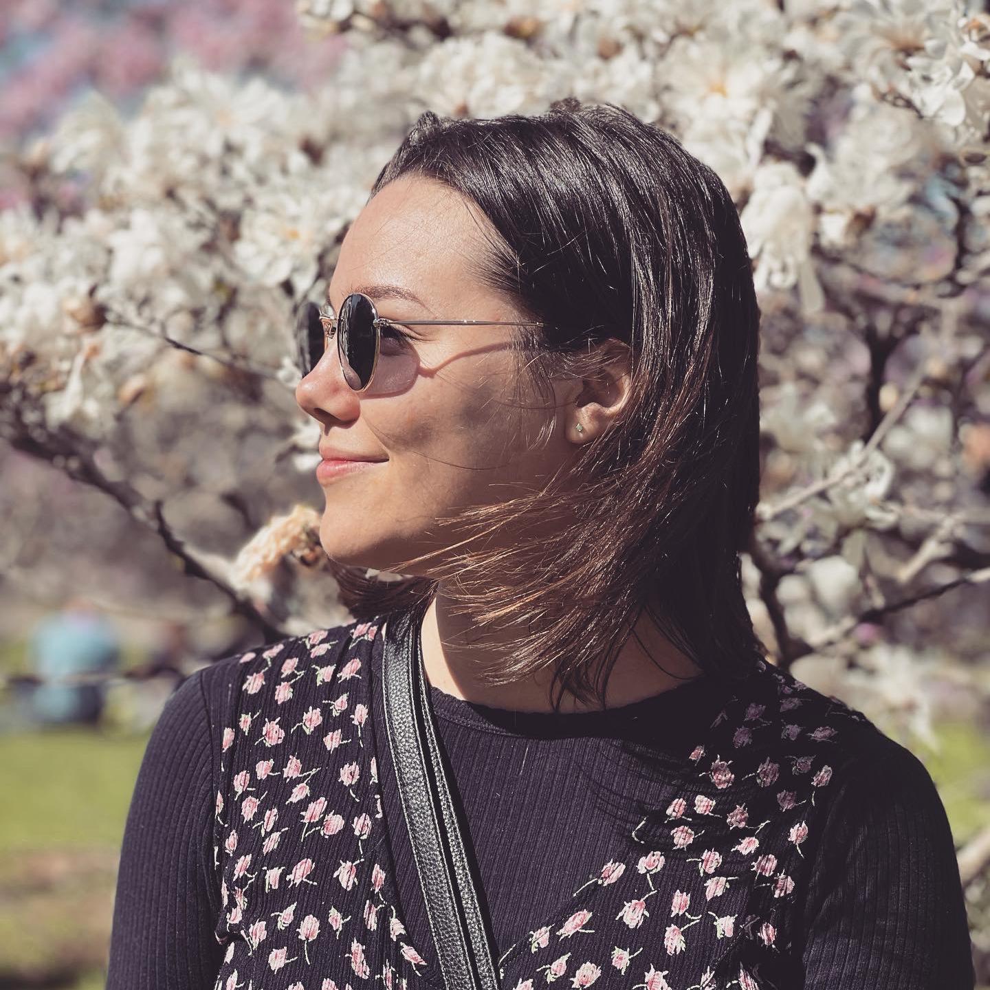 Image d'une femme au cheveux bruns portants des lunettes de soleil et une robe fleurie devant un arbre en fleur blanches